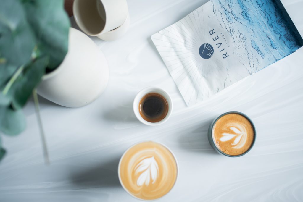 River Coffee Roasters bag of coffee next to 2 cups of milky coffee on a white table