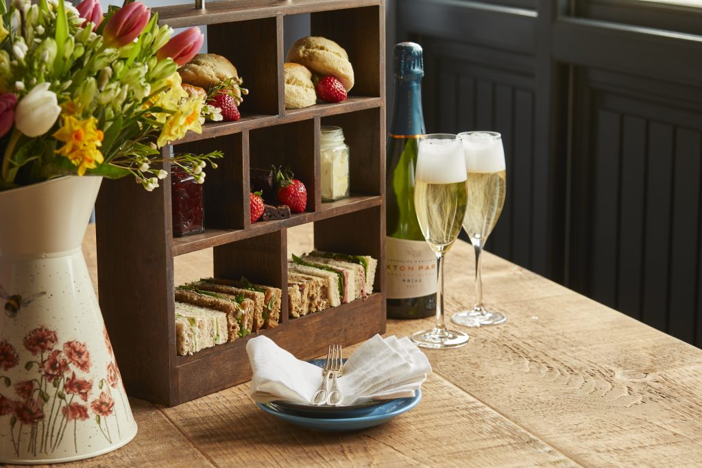 sandwiches and scones in a wooden frame stack with 2 glasses of sparkling wine and flowers