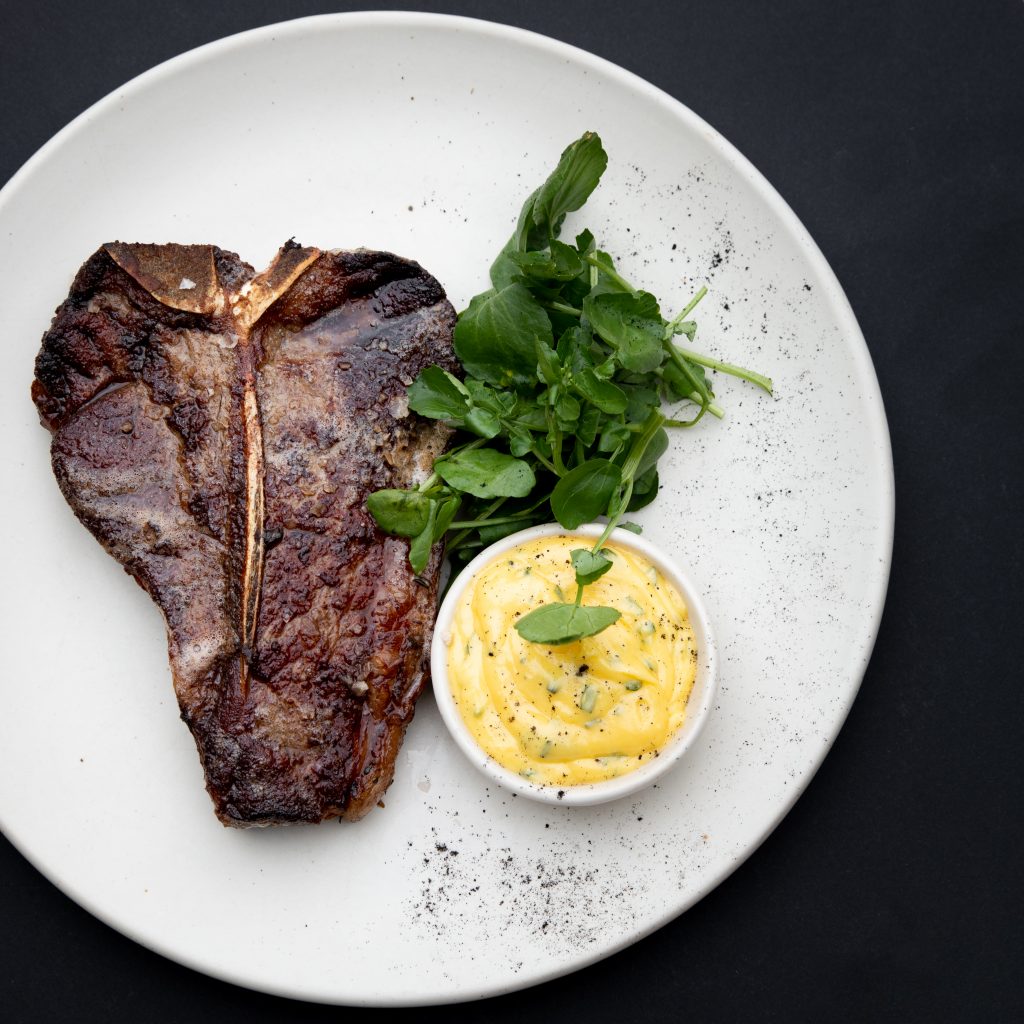 A plate with a T bone steak, watercress and bernaise sauce with vegetables
