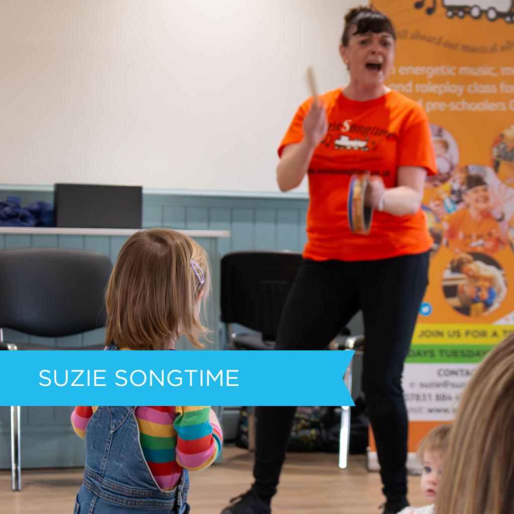 A woman leading a lively Suzie Songtime music session, playing a tambourine in front of young children, including a toddler in a rainbow-striped top and denim dungarees.
